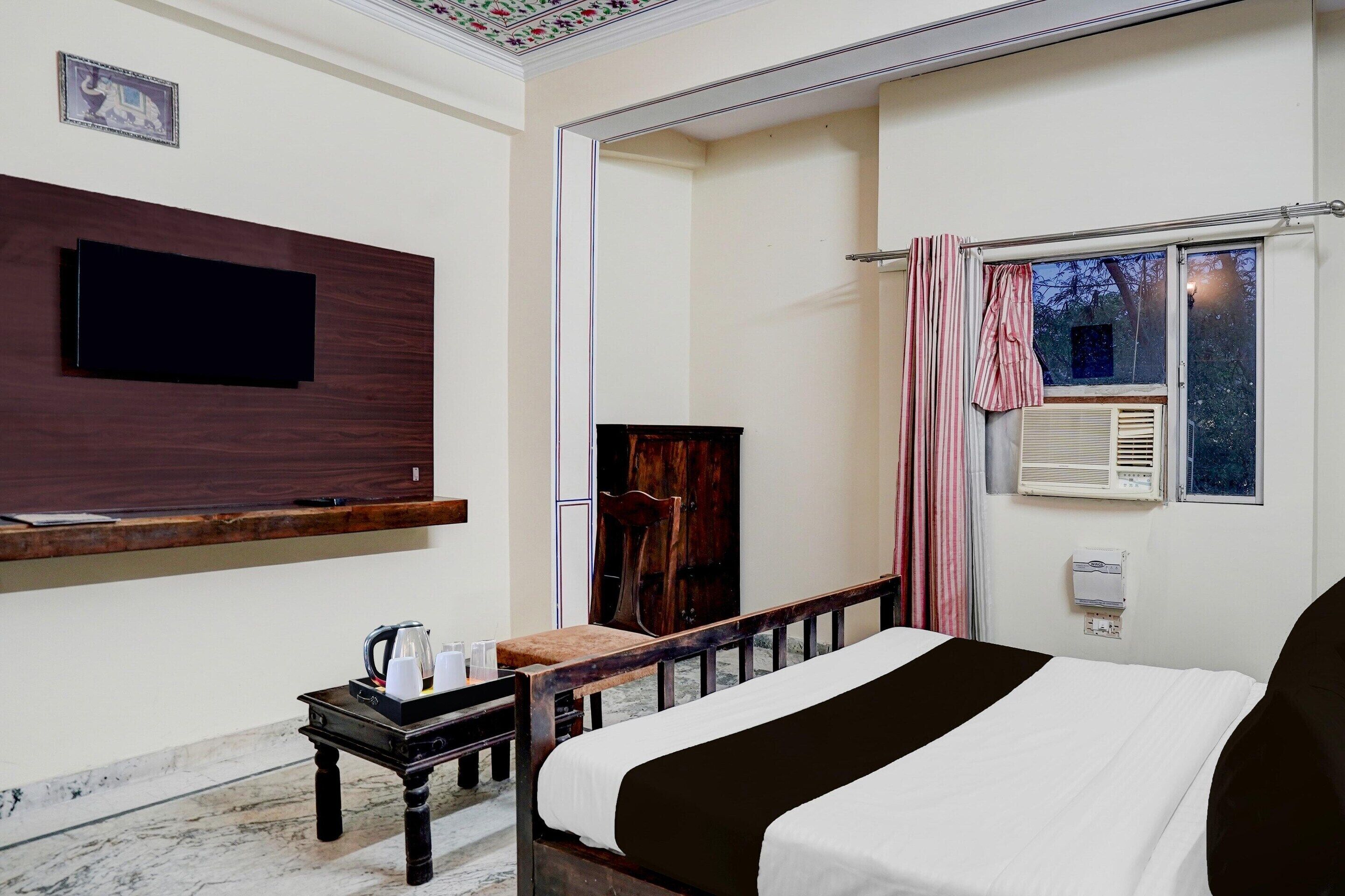 Habitación de hotel con cama, televisión de pantalla plana montada en la pared, una mesita con hervidor y tazas, ventana con cortinas y aire acondicionado también en la pared.