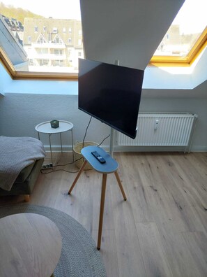 Ein Zimmer mit Fernseher auf einem Standfuß, einem kleinen runden Tisch, einer Couch und Blick auf Gebäude durch ein Oberlicht.