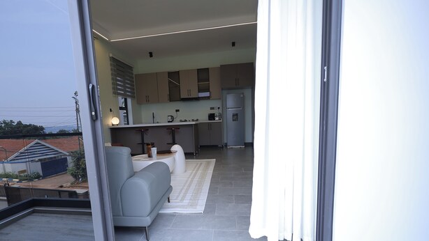 A modern kitchen with grey cabinets, a built-in oven, and a refrigerator. There is a grey sofa and a white rug in the living area. - Heritage Residences-Velvet Rouge 1BR (Kampala)