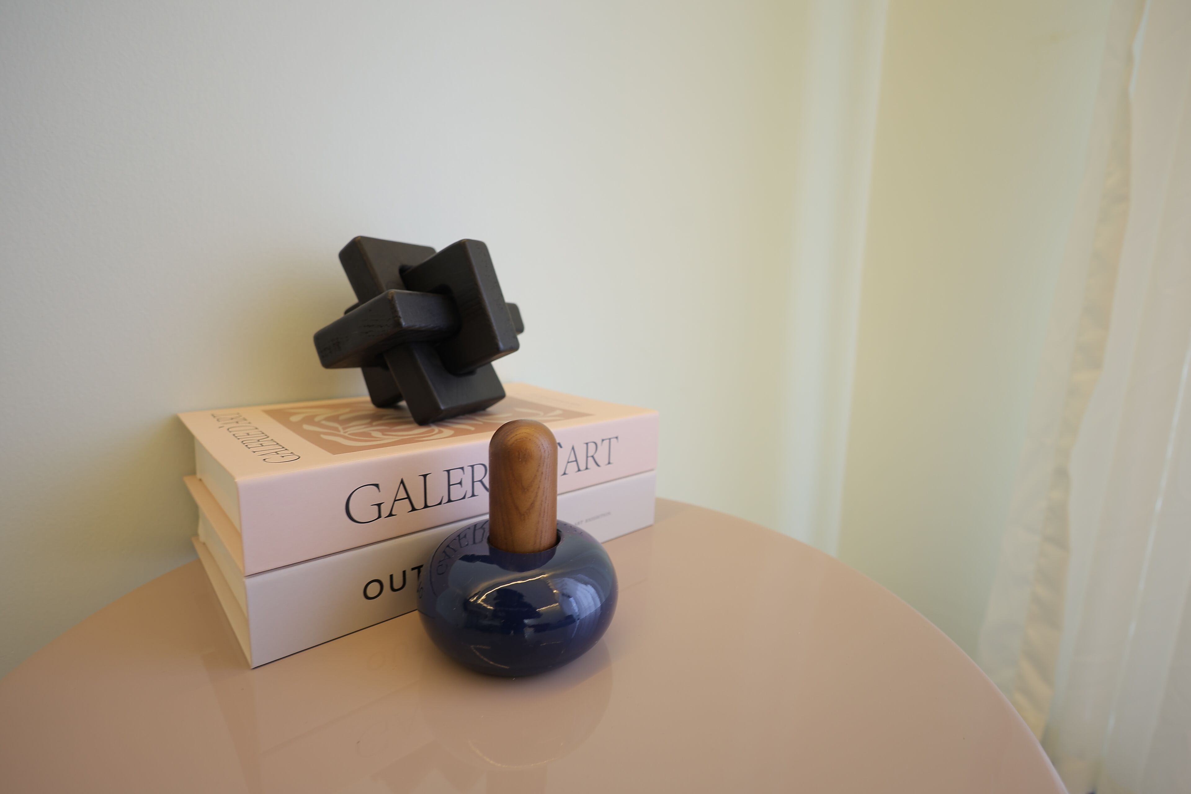A blue object on a table with a wooden handle, a white box with "GALERIA ART" text, and a white curtain in the background.