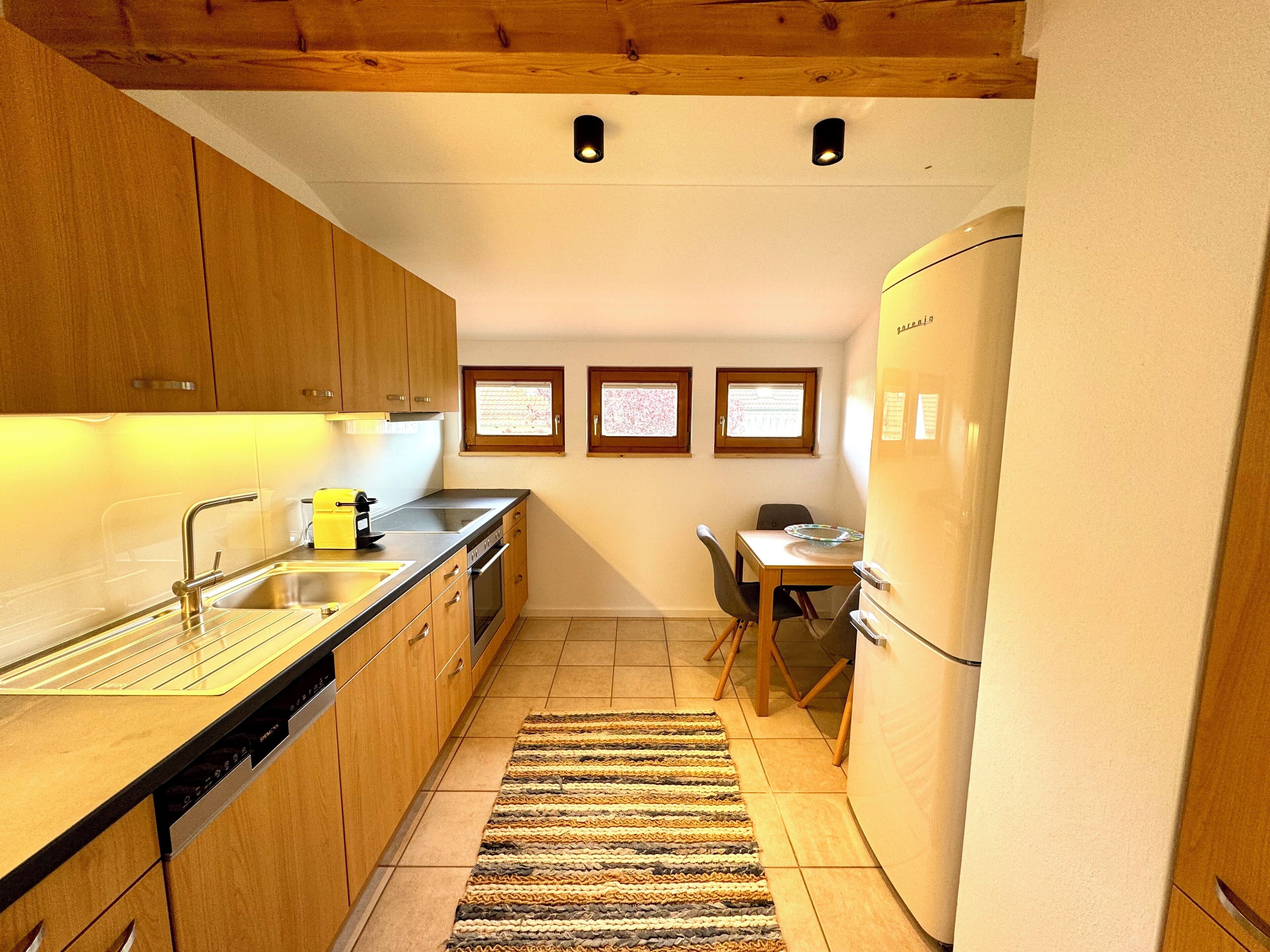 A compact kitchen with wooden cabinets, a built-in dishwasher, a sink, and a refrigerator. There are three framed pictures on the wall and a small dining table with chairs.
