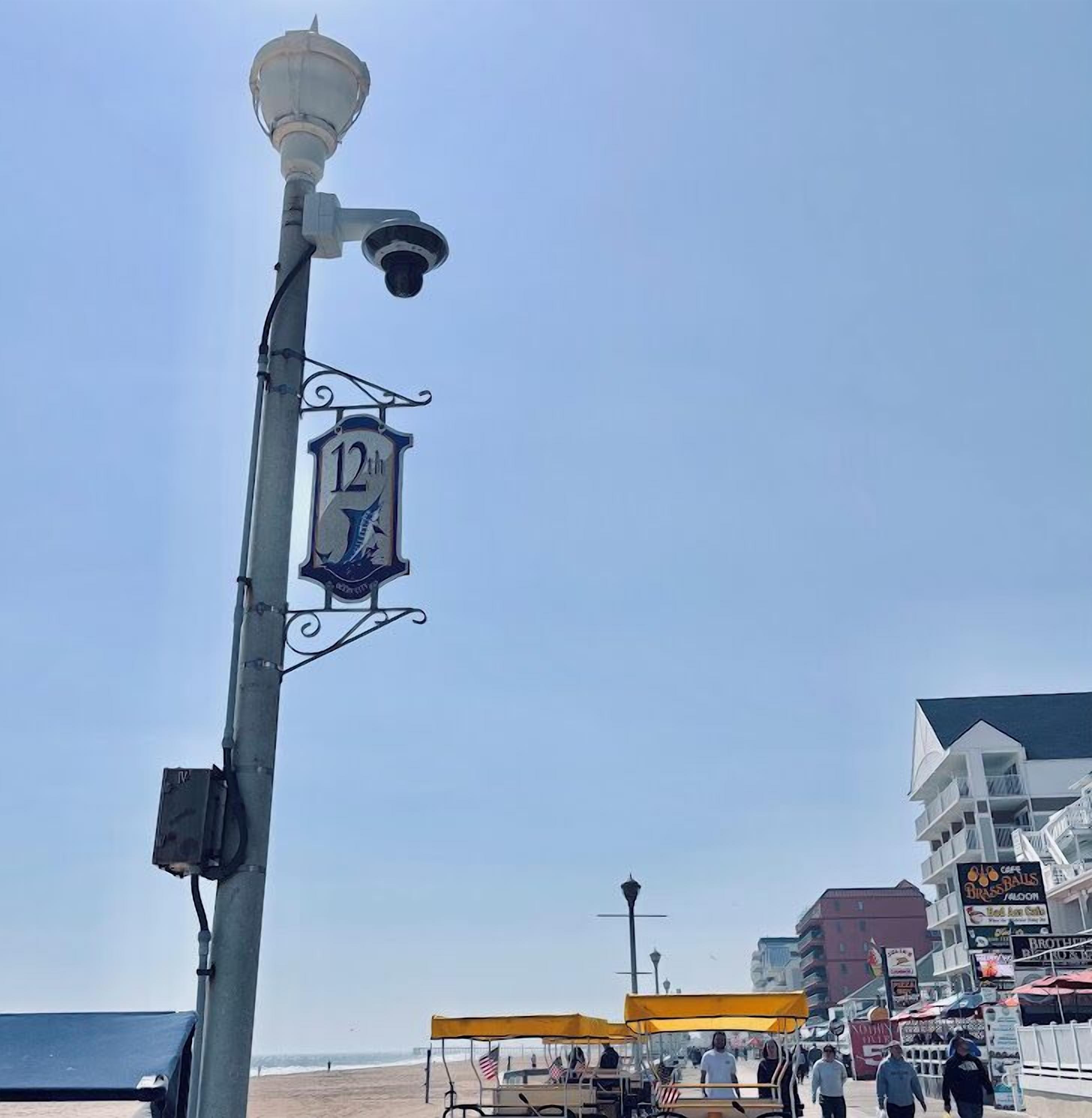 Un lampadaire avec une pancarte indiquant le 12e lieu, une plage avec des personnes et des chaises de plage, et des bâtiments avec des panneaux dans l’arrière-plan.