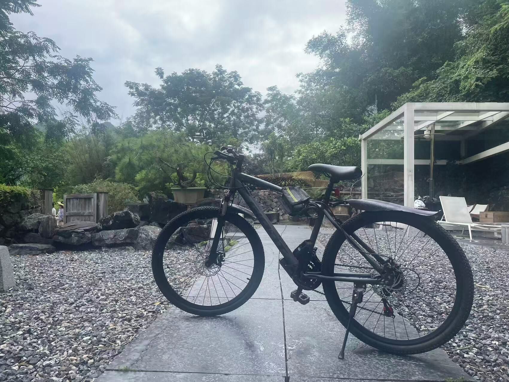 A bicycle parked in a garden with a stone pathway, a glass structure, and a wooden fence in the background.