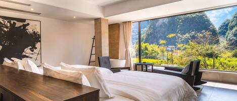 A modern hotel room with a large bed, a wooden desk, a chair, a small table, and a view of a mountainous landscape.