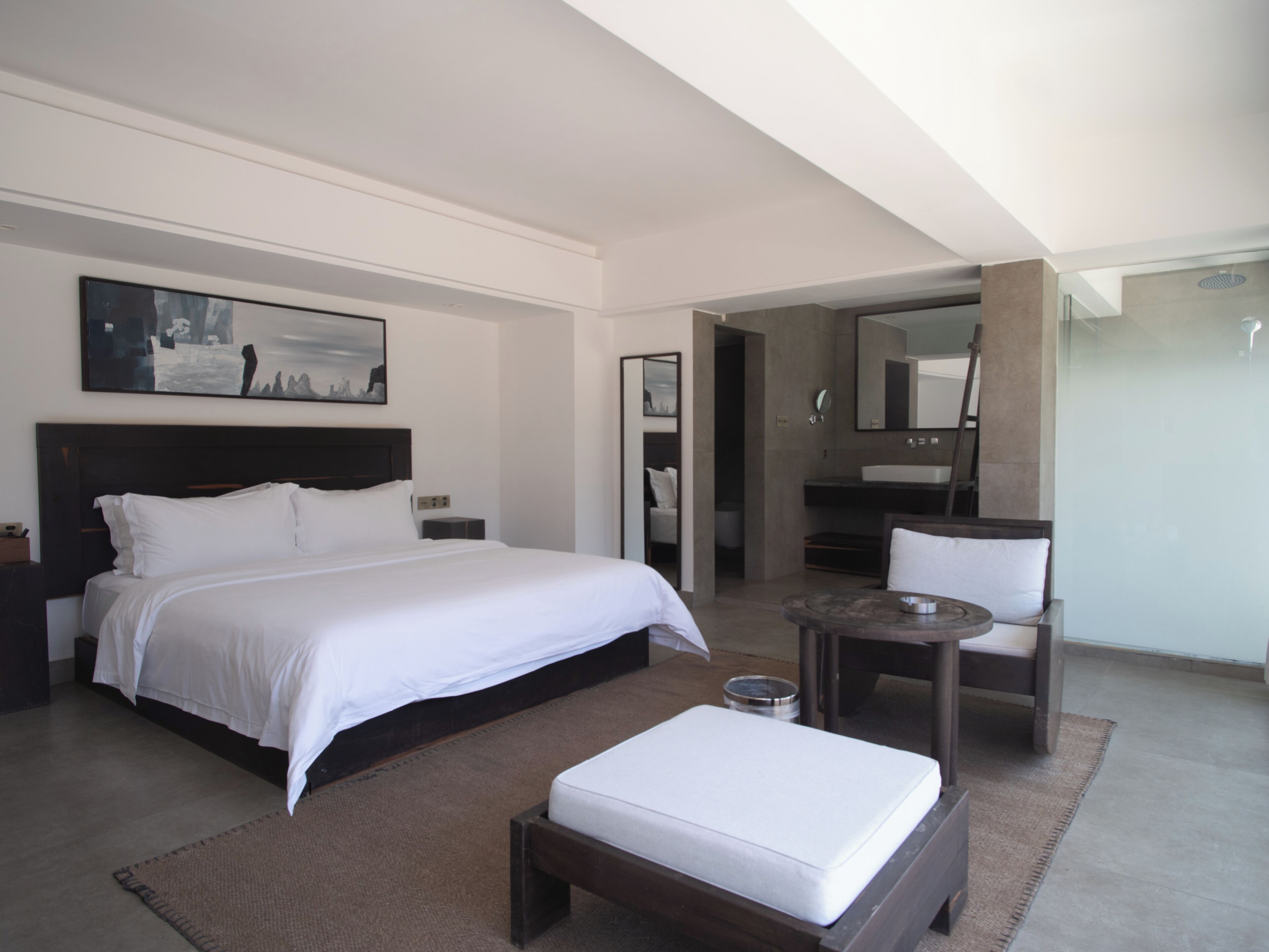 Una habitación de hotel moderna con una cama grande, una mesita redonda pequeña, una silla, un baño con espejo y un cuadro en la pared.