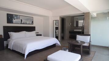 Una habitación de hotel moderna con una cama grande, una mesita redonda pequeña, una silla, un baño con espejo y un cuadro en la pared.