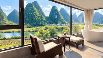 Une chambre d’hôtel moderne dotée d’une grande fenêtre offrant une vue sur un paysage montagneux et une rivière. On y trouve un canapé, une table avec un service à thé et une baignoire sur pieds.