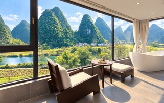 Habitación de hotel moderna con un ventanal que ofrece vistas a un paisaje montañoso y un río. Hay un sofá, una mesa con un juego de té y una bañera independiente.
