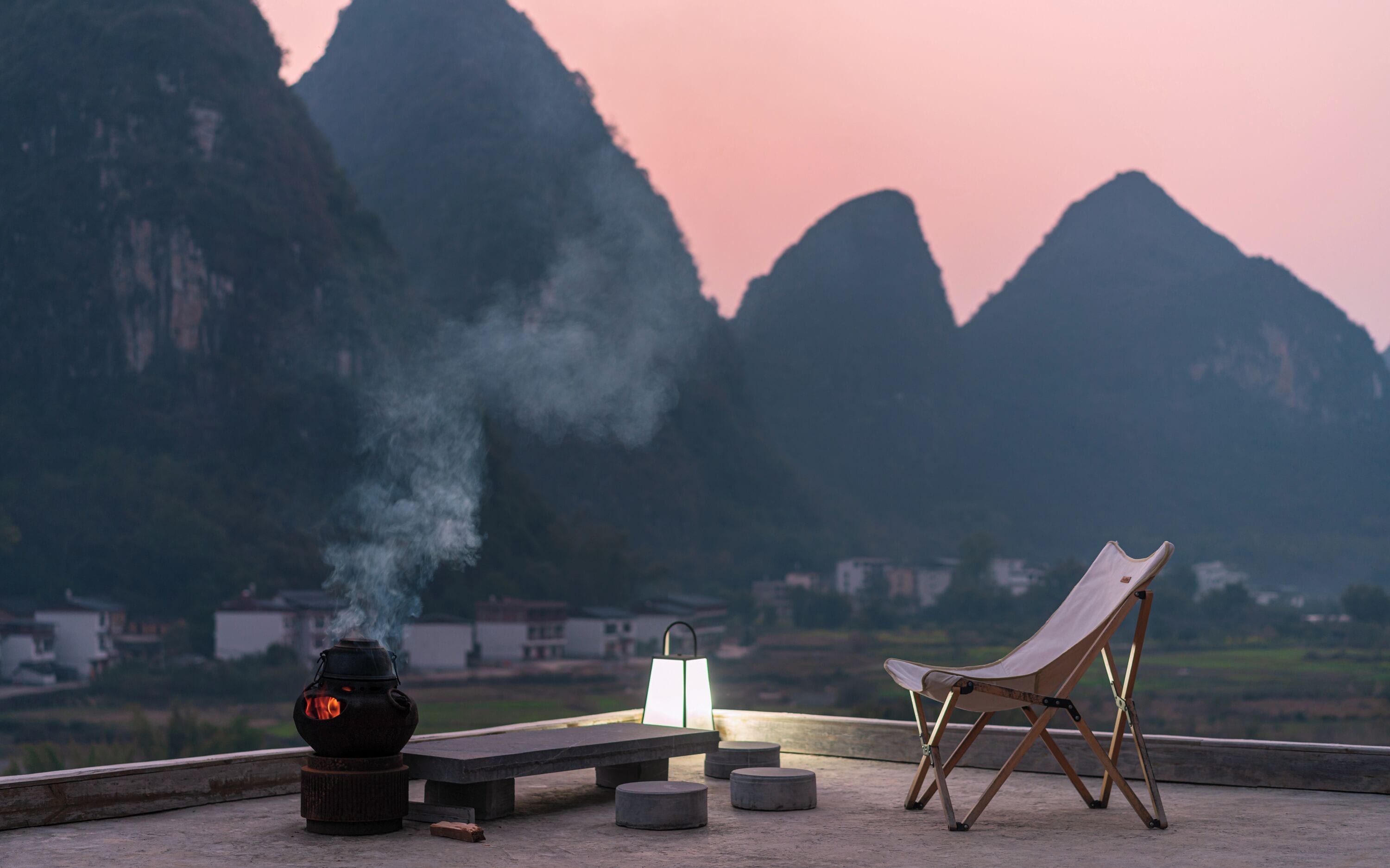 A table with a lantern and a chair on a rooftop overlooking a mountainous landscape.