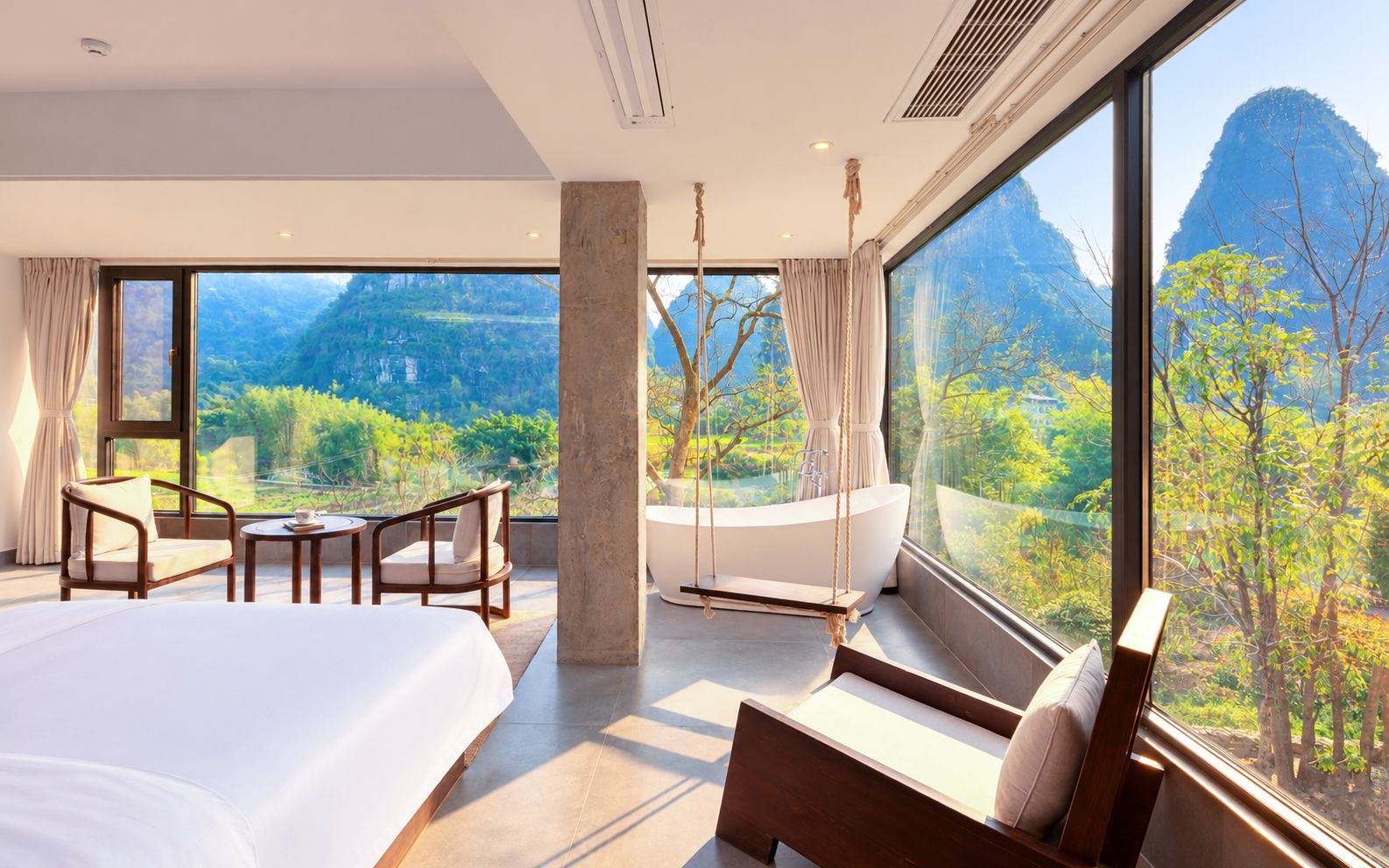Habitación de hotel moderna con una cama grande, un área de descanso con sillas, una bañera y vistas a un paisaje montañoso.