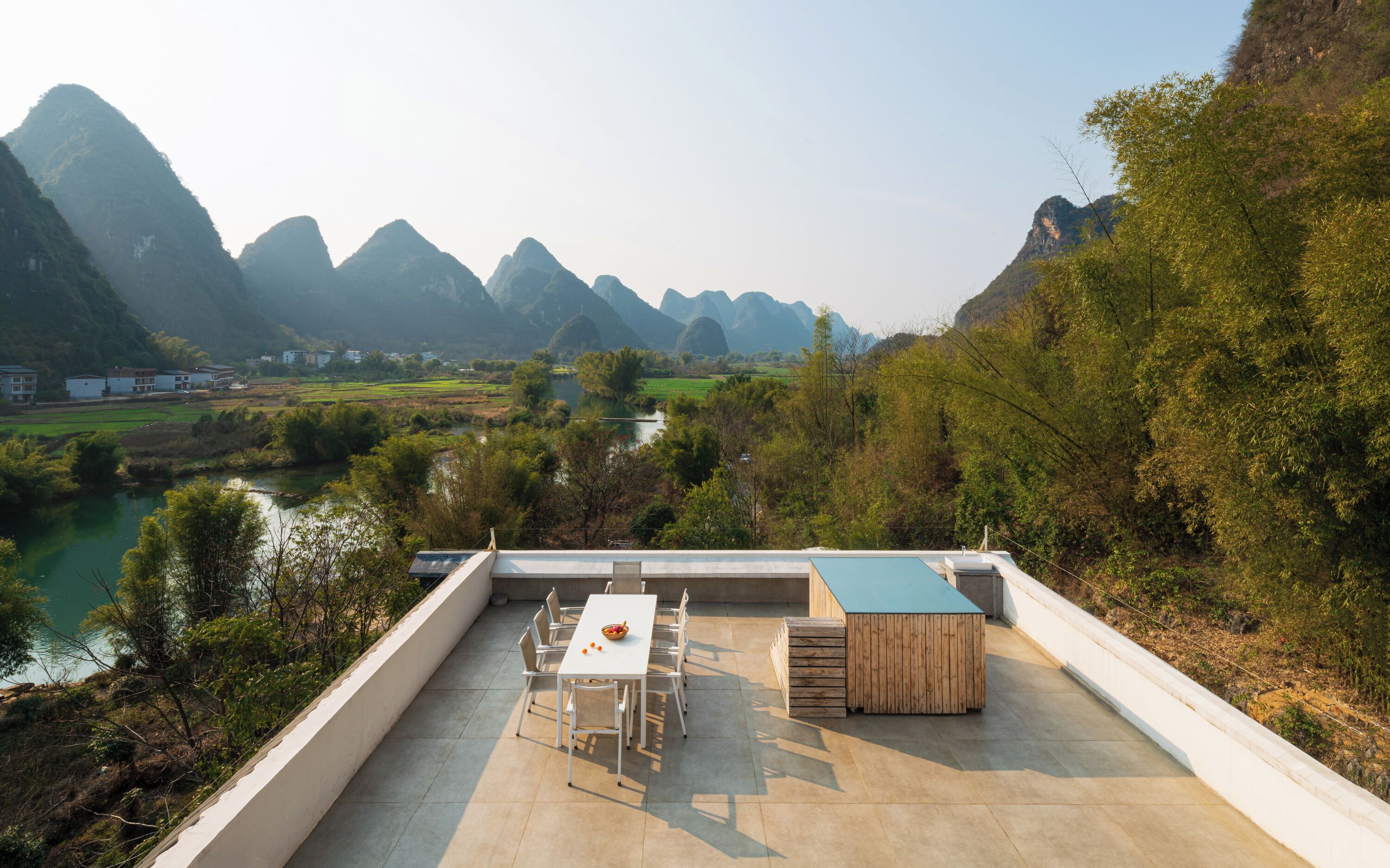 A rooftop terrace with a table and chairs, overlooking a river and mountains.