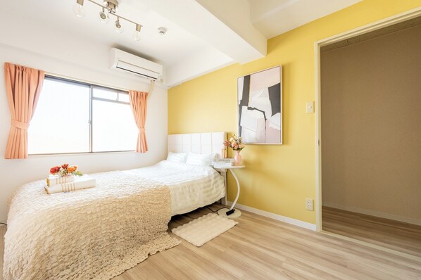 A bedroom with a bed, a bedside table with a vase of flowers, a window with curtains, and a wall-mounted air conditioner. - Akitsu 703 Kyocera Dome 10-minute walk to Dotonbori, Kansai Airport, Osaka Castle, Kaiyukan, USJ 1 transfer Family-friendly Free parking 15 (大阪府大阪市西区境川1丁目)