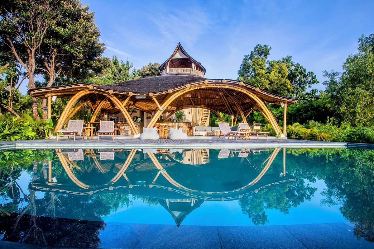 A poolside lounge with a wooden gazebo, lounge chairs, and a clear pool reflecting the surrounding trees.