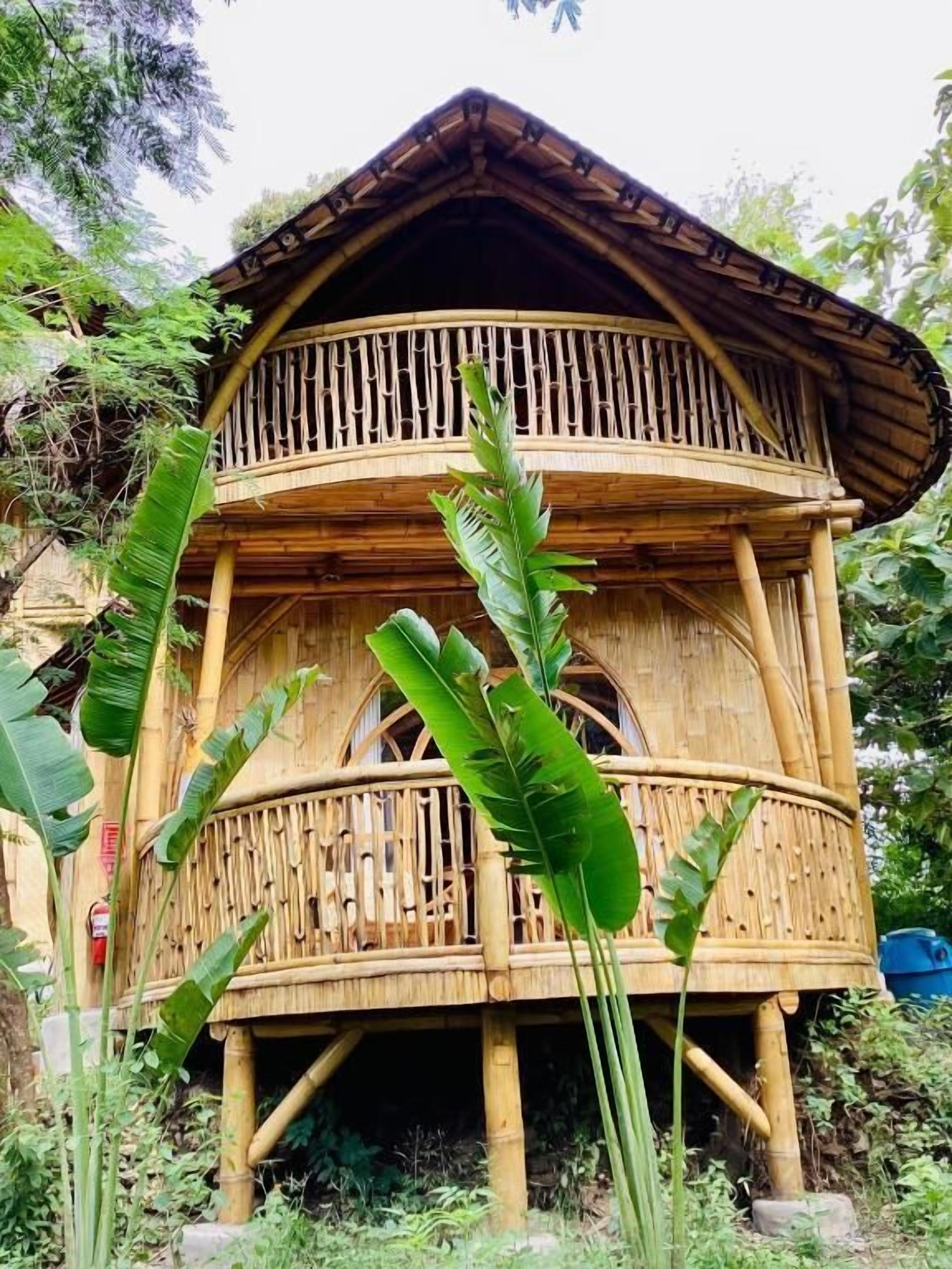 A two-story bamboo house with a balcony on the upper floor and a small entrance at the base.