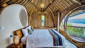 A bed with a white sheet, a wooden headboard, and a bedside lamp. There is a window with a view of greenery and a body of water.