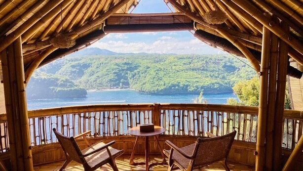 A wooden deck with a table and chairs, overlooking a body of water and green hills.