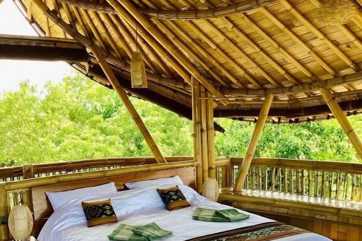A bed with white linens and a patterned blanket, a wooden headboard, and a view of green foliage through a thatched roof structure.