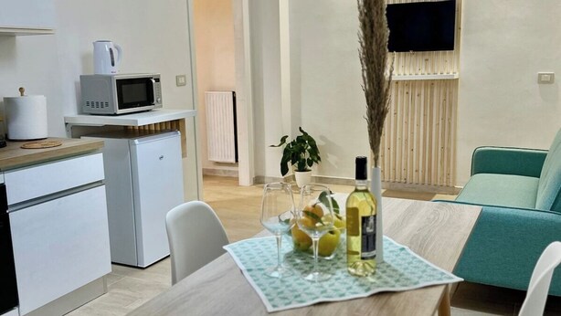 Una cocina moderna equipada con microondas, cafetera y un refrigerador pequeño. Se observa una mesa de comedor con un mantel individual, una botella de vino y una planta. También se ve un sofá color turquesa y un televisor montado en la pared.