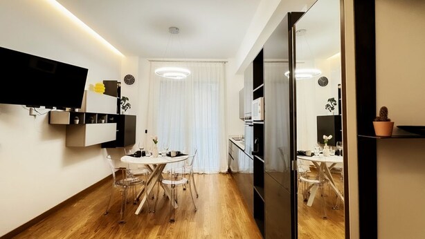 Uma cozinha moderna com forno embutido, sala de jantar com mesa redonda e cadeiras, e um espelho grande que reflete o espaço.