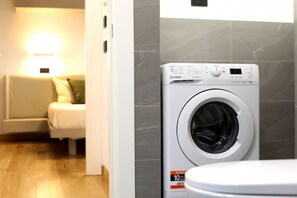 A modern laundry room with a front-loading washing machine, a sofa, and a wall-mounted light. - Hostmate - Milano - Pistrucci  (Milano)