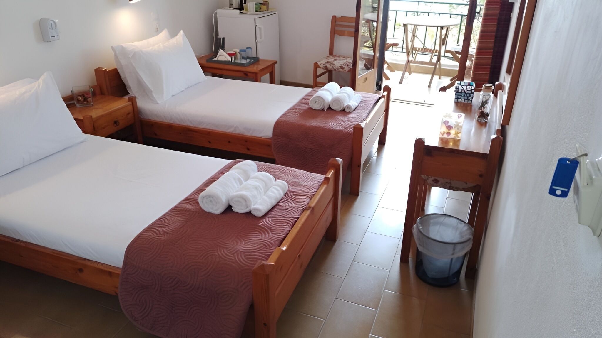Habitación de hotel con dos camas, cada una con ropa blanca y toallas enrolladas. Hay una mesita de noche de madera con una lámpara, una pequeña mesa con una silla y vistas al balcón exterior.