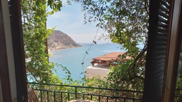 A balcony with a table and chairs, overlooking a coastal view with a mountain and a building. - Loggia (Agia Galini)