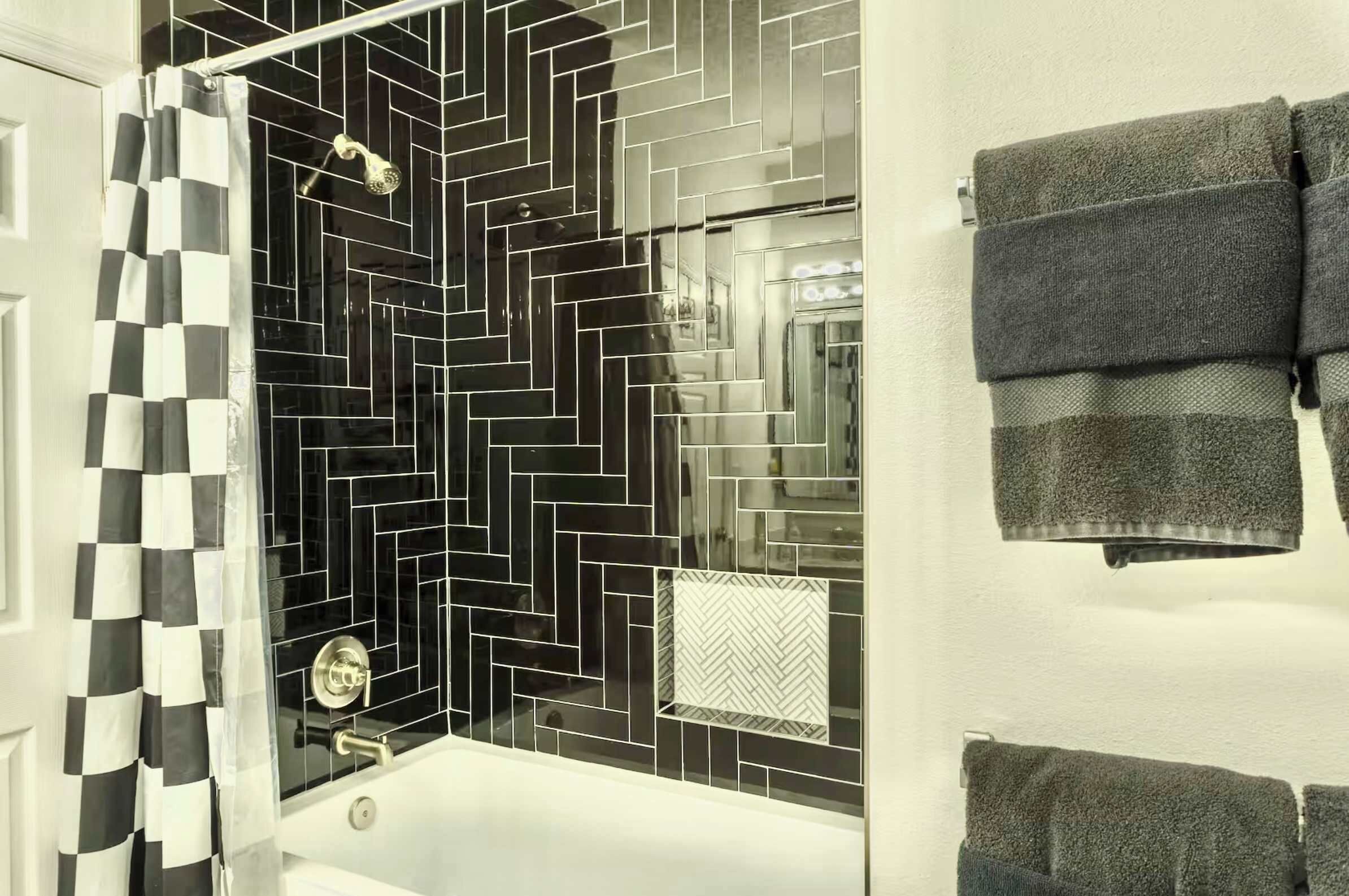 Un baño con cortina de ducha a cuadros blancos y negros, salpicadero de azulejos en espiga blanco y negro, y un perchero con toallas dobladas.