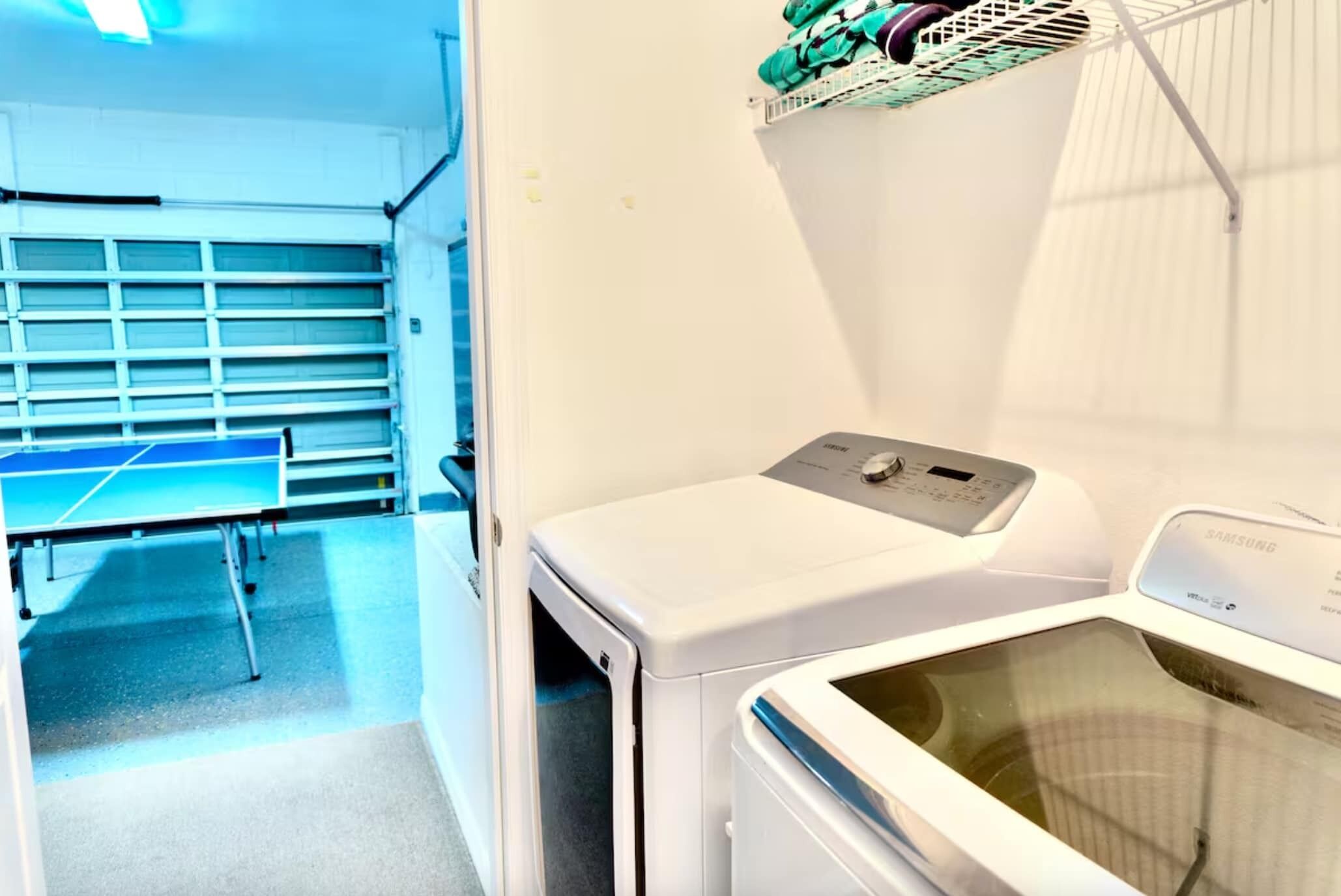 Un área de lavandería con lavadora y secadora, una mesa de ping pong y un estante con ropa doblada.