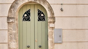Une porte à deux battants, peinte en vert et ornée d’éléments décoratifs, un arc en pierre et une petite fenêtre avec des volets.