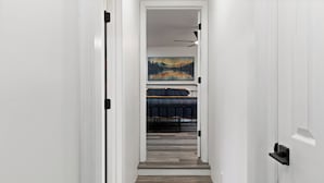 Un couloir étroit, doté d’un plancher en bois, mène à une pièce où se trouvent un lit et un tableau accroché au mur.