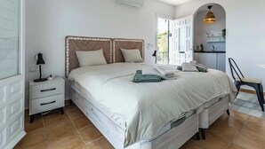 Une chambre à coucher comprenant un lit, une table de chevet, une lampe et une porte ouverte donnant sur un espace cuisine.