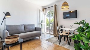 Un salon moderne avec un canapé gris, une table à manger avec des chaises, un téléviseur fixé au mur et une grande fenêtre avec des rideaux.