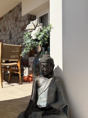 Una estatua de una figura sentada, posiblemente Budha, frente a una pared con otra pared de piedra y una silla al fondo.
