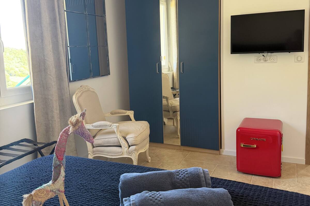 A hotel room with a bed, a chair, a TV, a red suitcase, and a mirror.