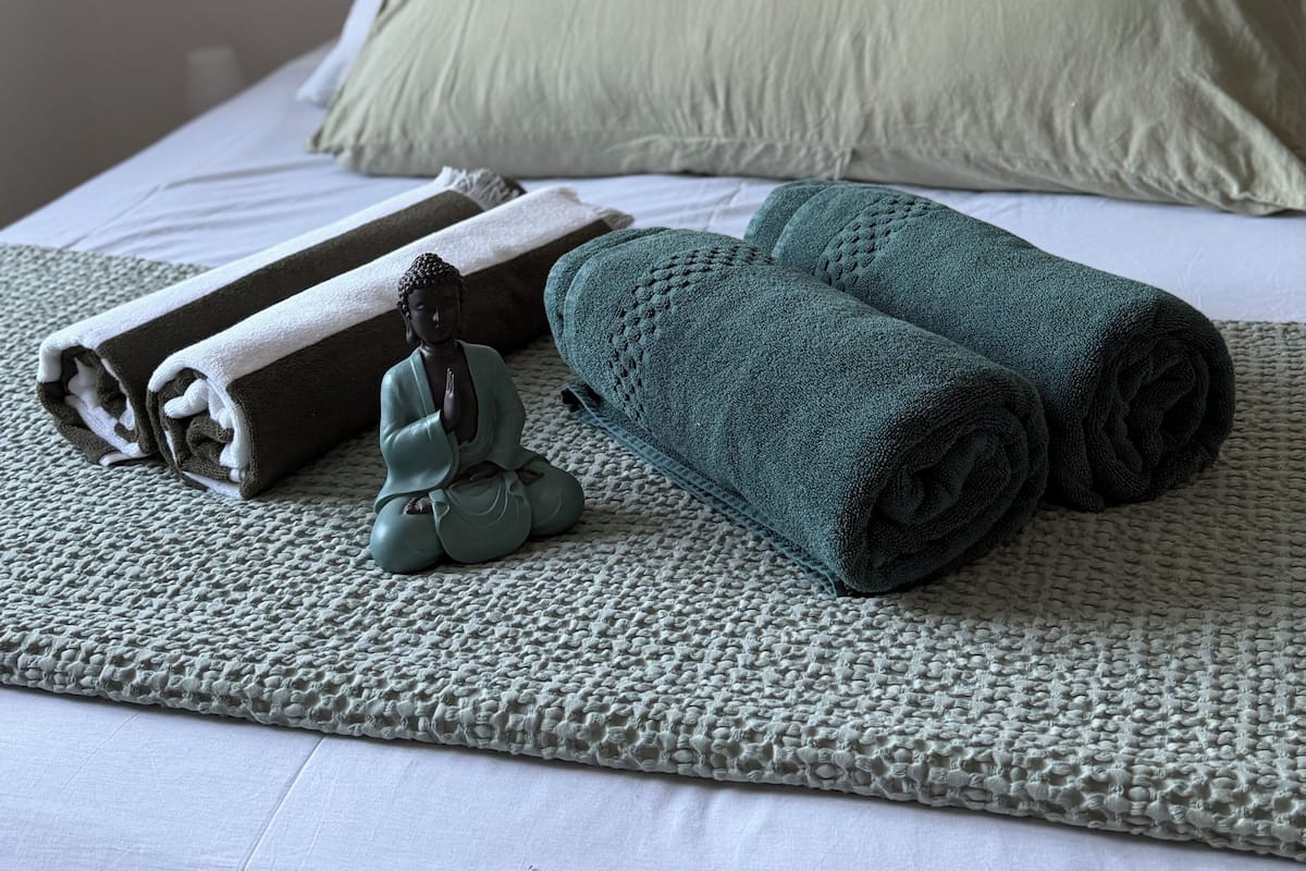 A neatly made bed with white linens, two rolled towels, a small Buddha statue, and a dreamcatcher on a textured grey blanket.