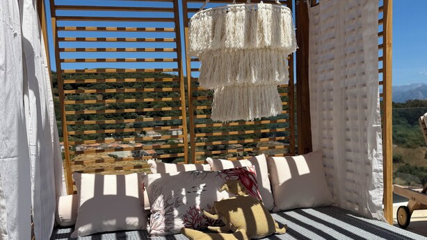 A wooden pergola with a hanging macrame piece, a cushioned seating area with patterned cushions, and a view of a mountainous landscape. - Petit Coin de Paradis (Alata)