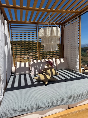 Una pérgola de madera con un adorno colgante de macramé, una zona de estar con cojines estampados y vistas a un paisaje montañoso.