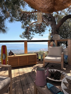 A wooden deck with a view of the city, a surfboard, a chair, and a table with pinecones. - Petit coin de paradis (Alata)