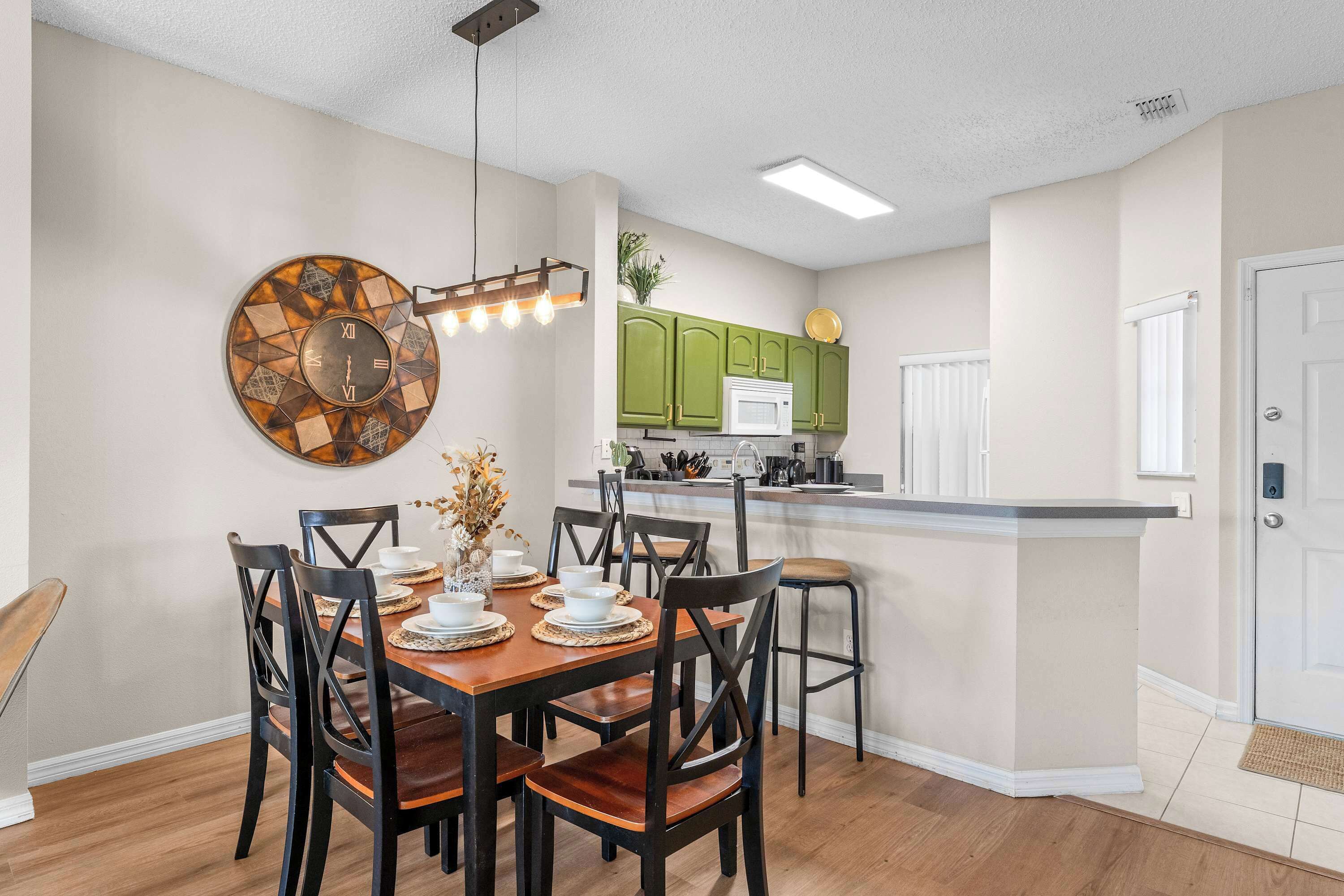 A dining area with a wooden table and chairs, a wall clock, and a kitchen area with green cabinets and a white countertop.