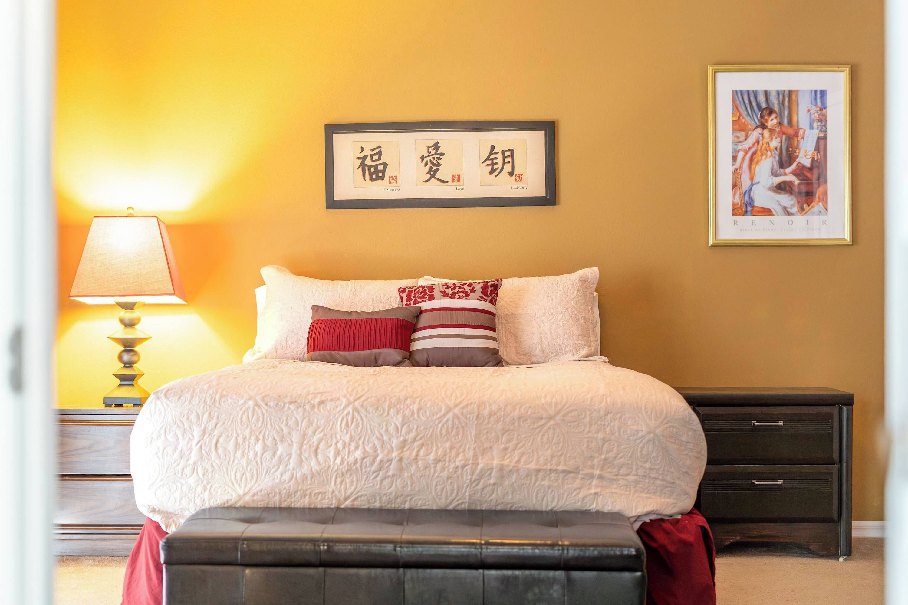 A neatly made bed with a patterned bedspread and multiple pillows, a bedside lamp, a framed artwork on the wall, and a dark nightstand with drawers.