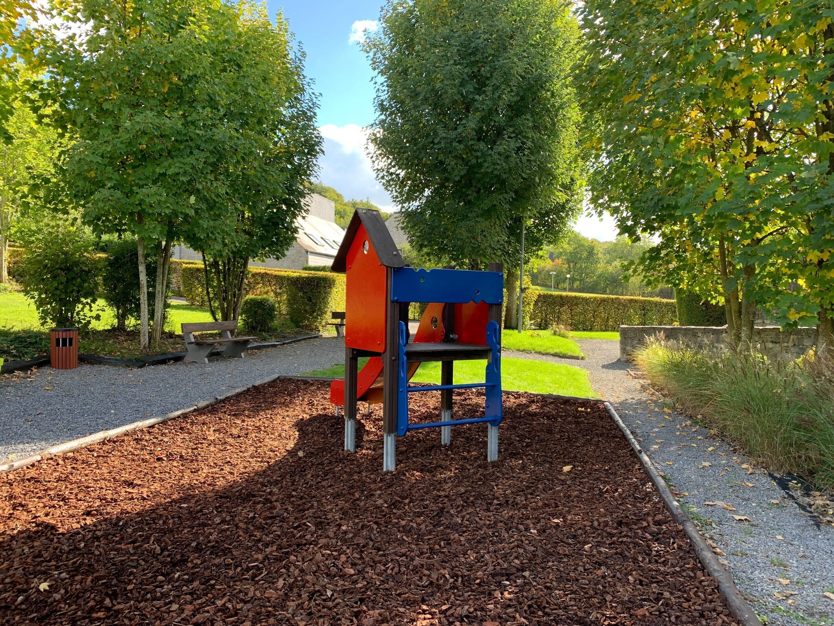 A playground with a red and blue play structure surrounded by mulch, trees, and a gravel path.