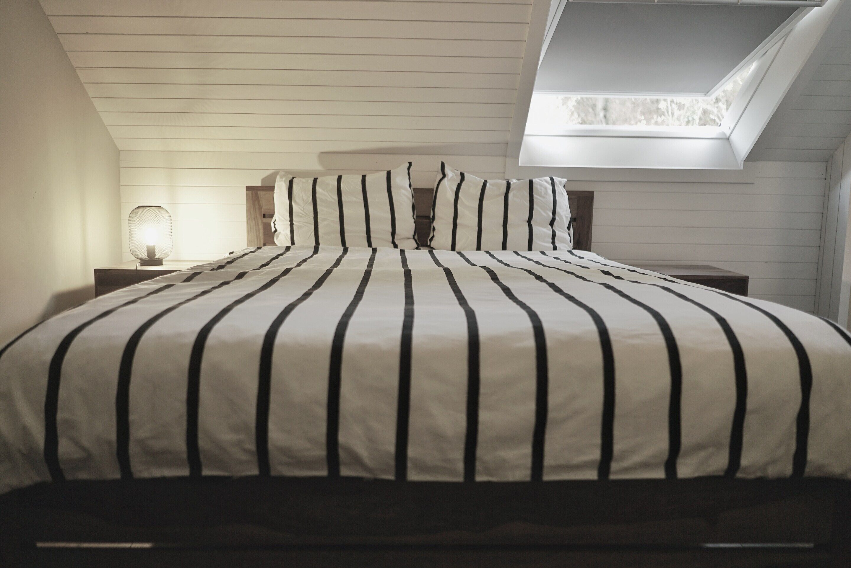 A bedroom with a sloped ceiling, a bed with a striped bedspread, a nightstand with a lamp, and a skylight.