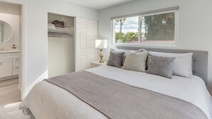 A bedroom with a large bed, a nightstand with a lamp, a window with a view of trees, and a door leading to a bathroom. - 2 Mi to Dtwn Palm Springs! Modern Condo w/ Pool (Palm Springs)