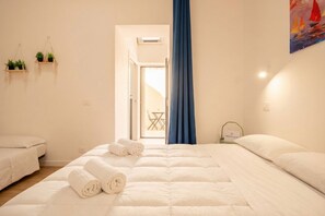 Un dormitorio con cama, dos mesitas de noche, ventana con cortinas, planta en la pared y puerta que da a otra habitación.