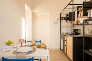 A dining area with a white table set for two, a kitchen with black shelving, and a door leading to another room.
