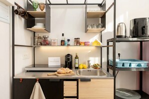 Una cocina compacta con un lavamanos, una cafetera y diversos utensilios de cocina en estantes.