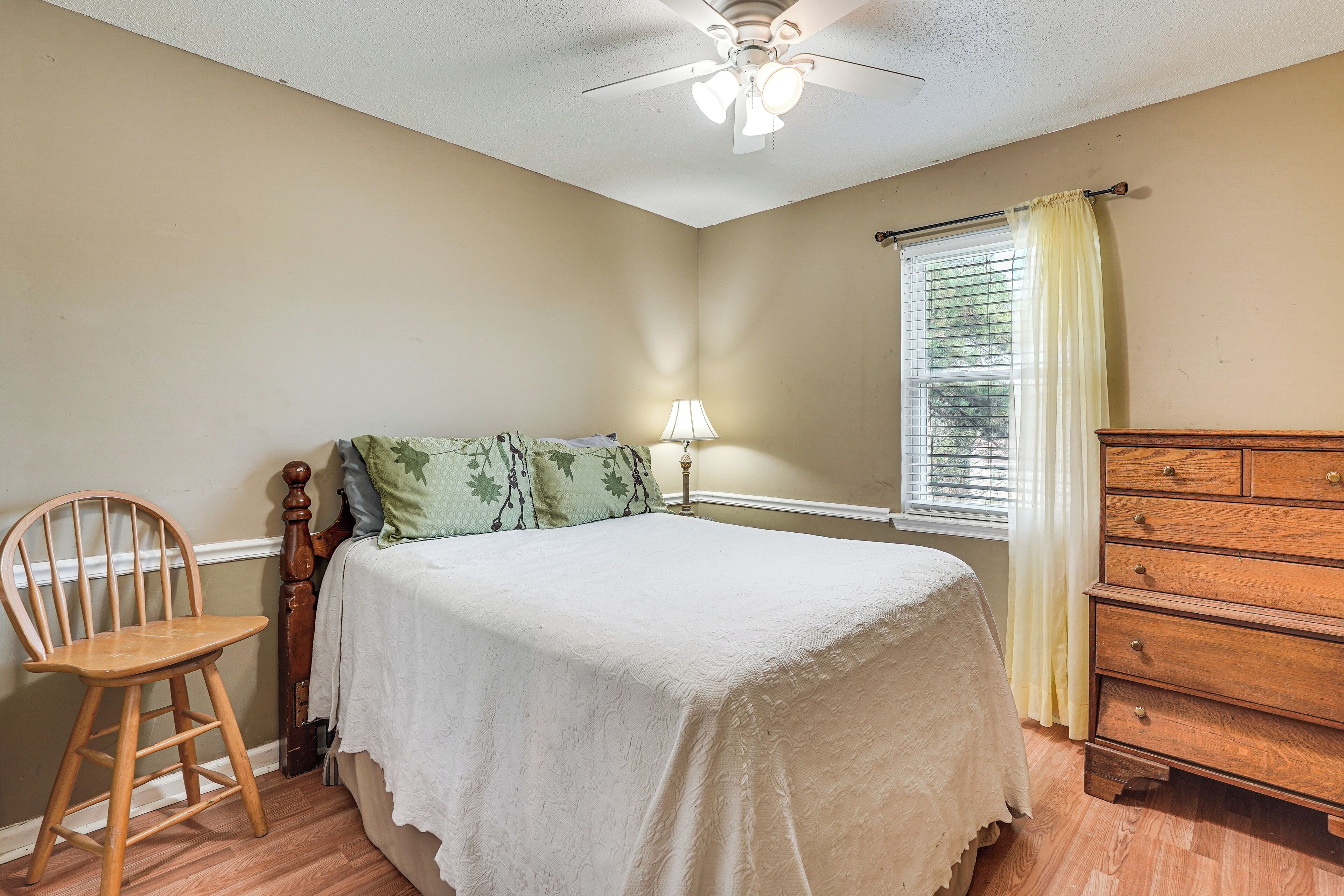 A bedroom with a bed, a wooden chair, a dresser, a ceiling fan, and a window with curtains.