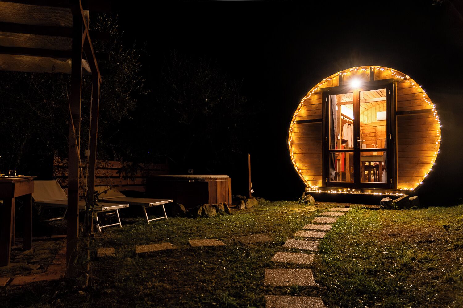 Eine runde Holzkonstruktion mit Glastür, beleuchtet von Lichterketten, und ein Kiesweg, der dorthin führt.