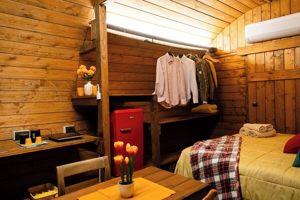 A cozy cabin room with wooden walls, a bed with a plaid blanket, a red heater, and a table with a vase of flowers.