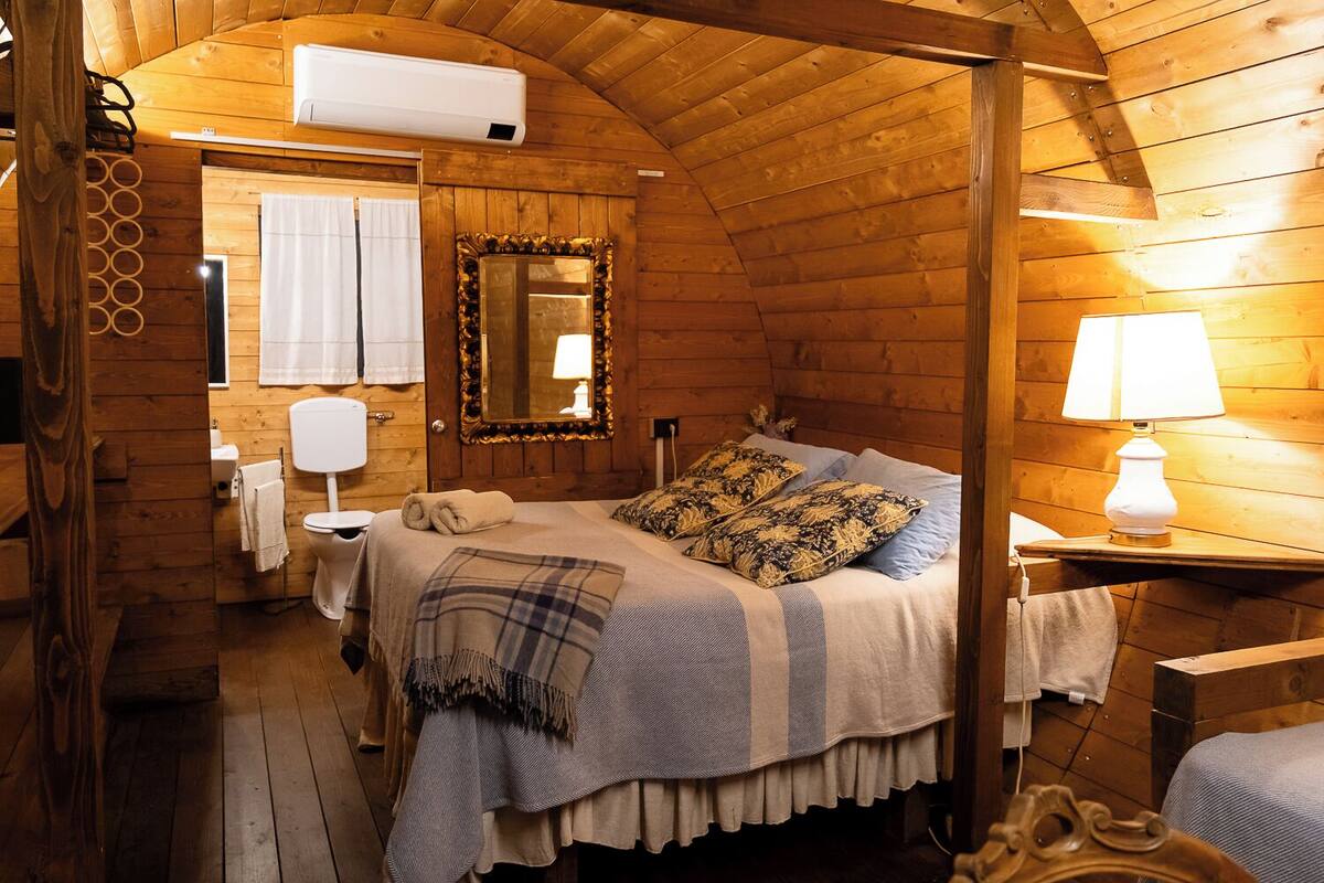 A cozy wooden cabin bedroom with a bed, a bedside lamp, a mirror, and a window with curtains.